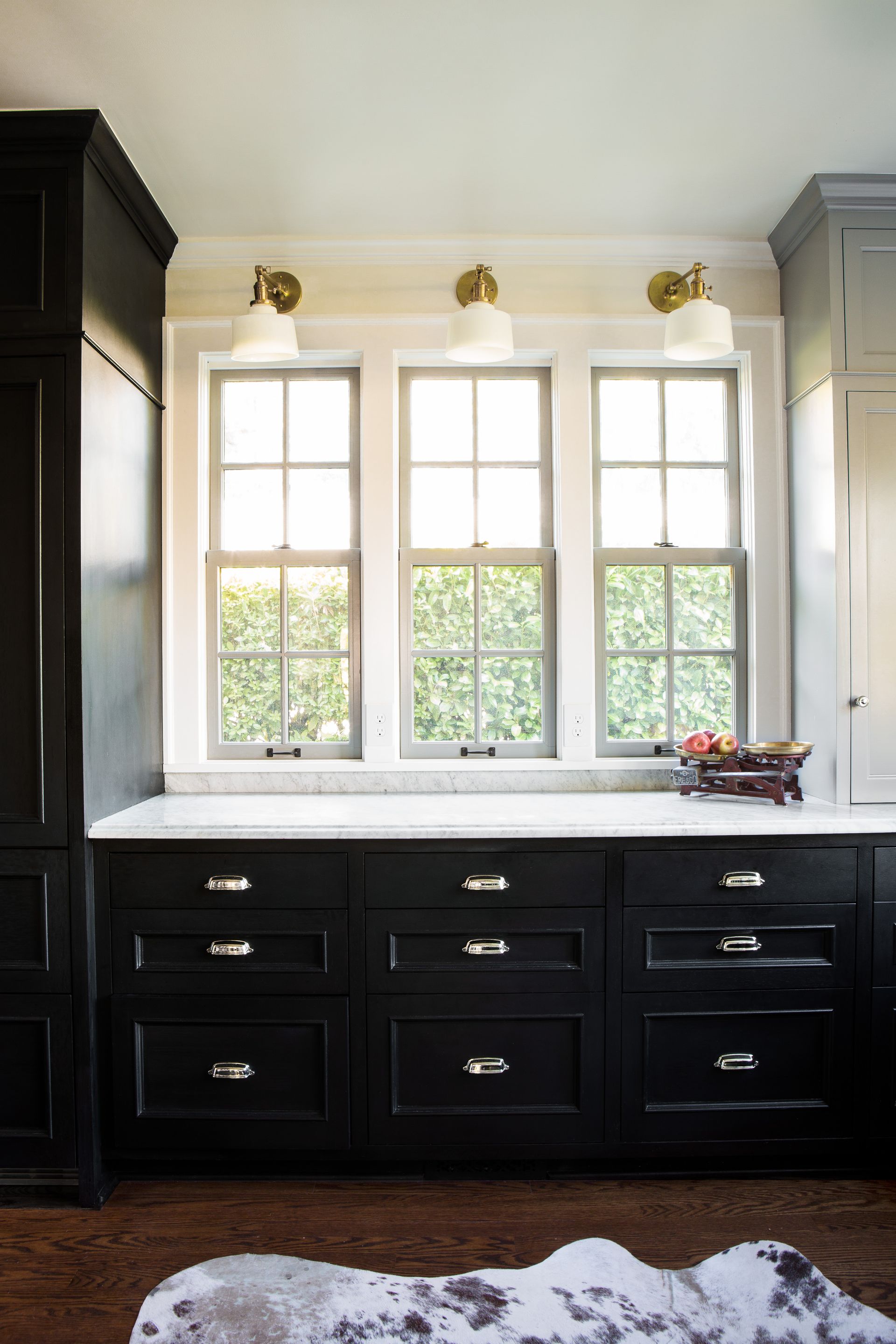 A trio of kitchen windows with brass sconces above and a bank of ebonized cabinets below.