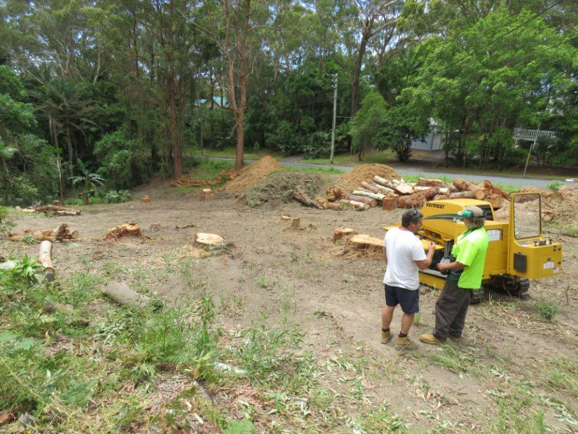 Two Men Are Standing in a Dirt Field Next to a Stump Grinder — Jeff Pearce Tree Services in Lansdowne, NSW