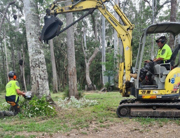 A Man is Sitting on the Ground Next to a Yellow Excavator — Jeff Pearce Tree Services in Tuncurry, NSW