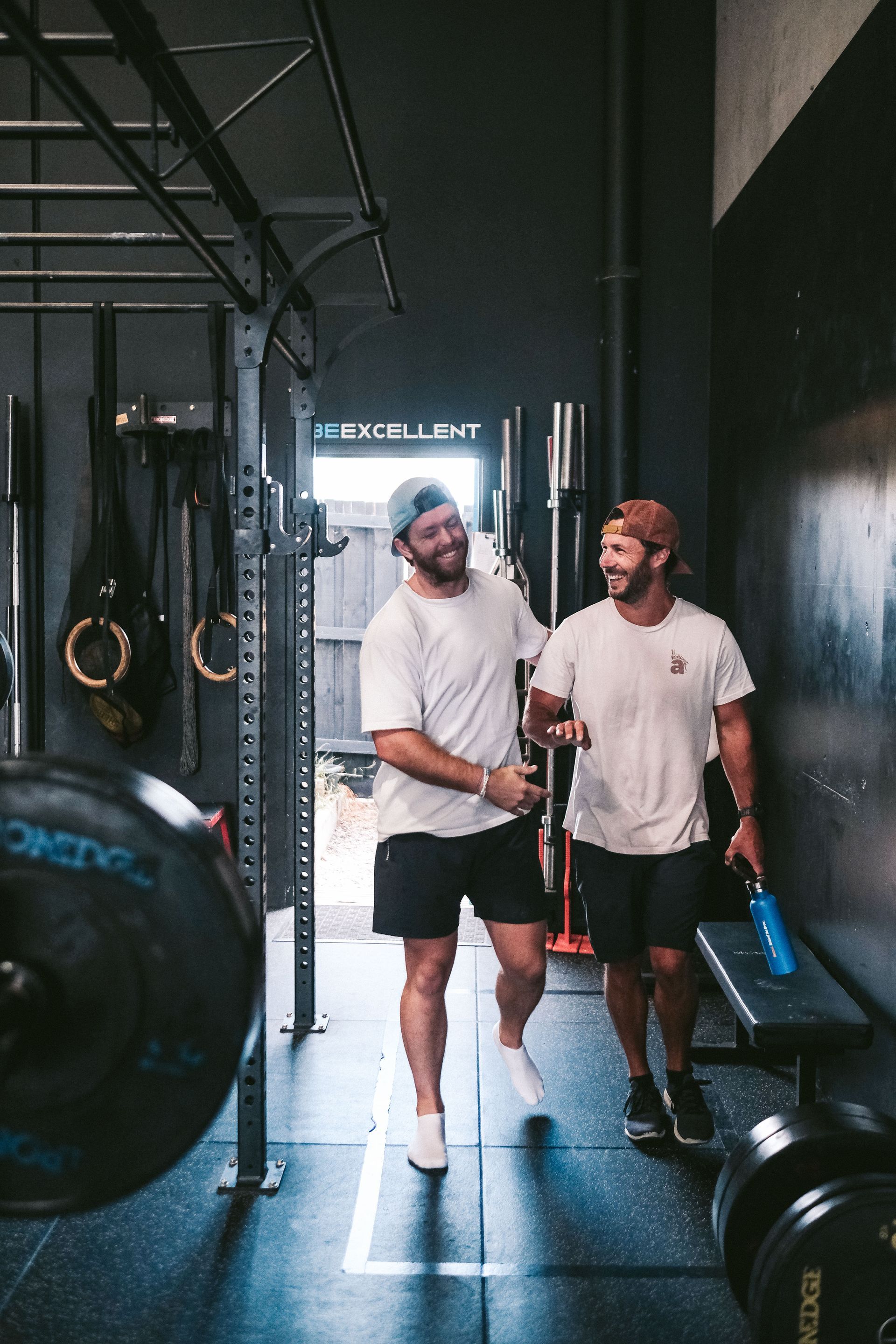 Two men in a gym smiling, one holding the other’s arm. Black shorts, white shirts and caps. Bench and weights nearby.