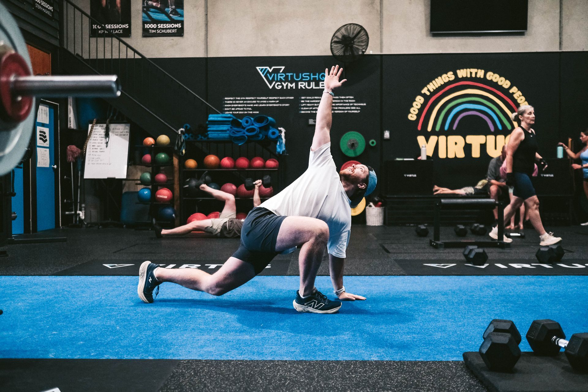 Man lunging, arm raised, in a gym with others exercising. Blue mats, weights, and a rainbow mural in the background.