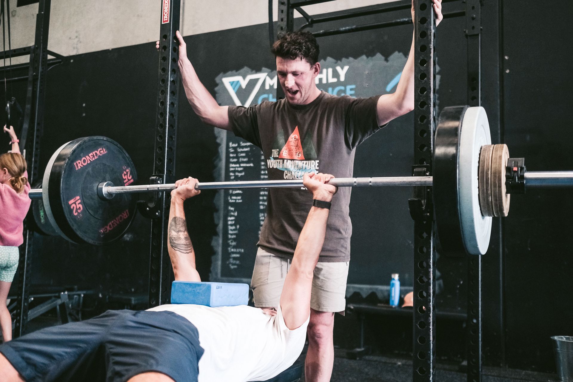 Man bench-pressing with spotter at a gym. Blue block under his chest. Black weights on bar.