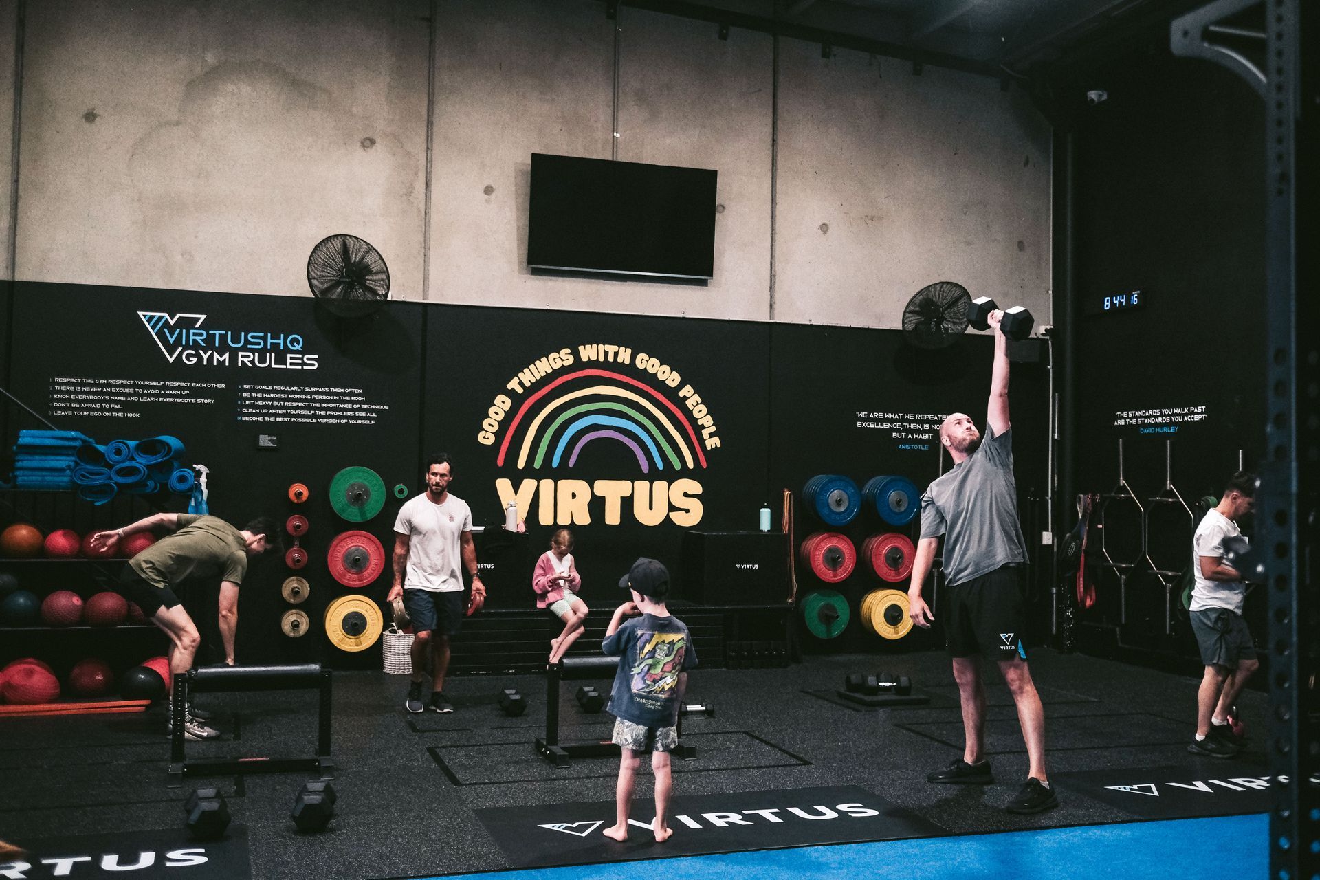 People exercising in a gym: lifting weights, jumping, and observing. A rainbow-themed wall with