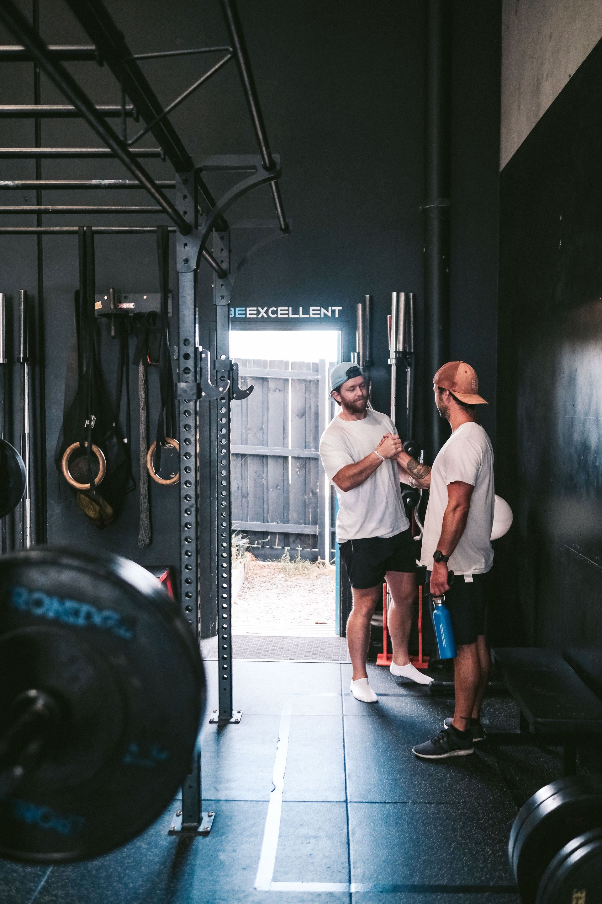 Two men in gym greeting; one in a cap, one in white tee. Dark interior, weight rack, and a doorway to outside.