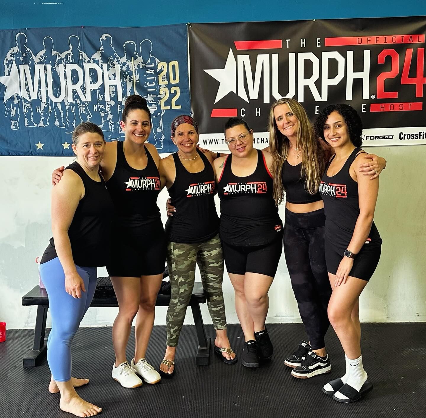 Group of six women in athletic wear pose, smiling, inside a gym. Two banners are visible in the background.