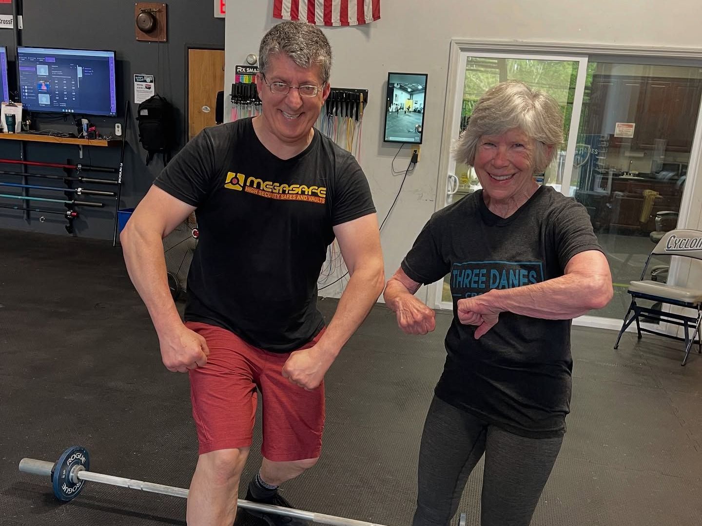 Man and woman flexing muscles at a gym. Man in red shorts, woman in gray pants, both smiling.