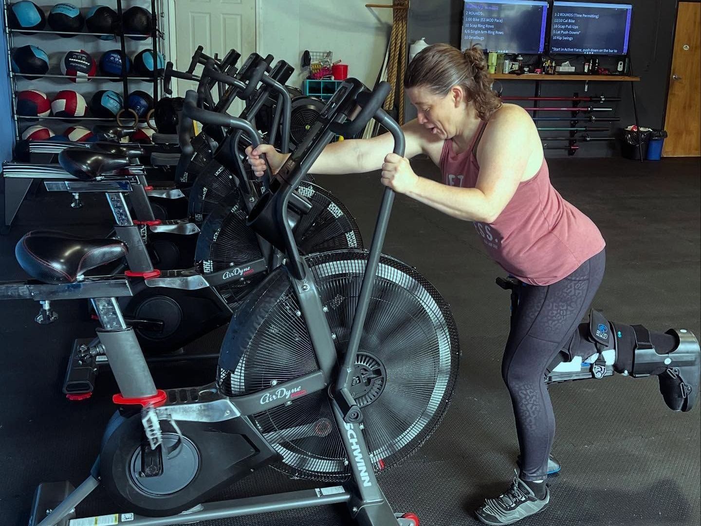 Woman exercising on a black air bike in a gym. She's wearing athletic clothing, arms pulling, face strained.