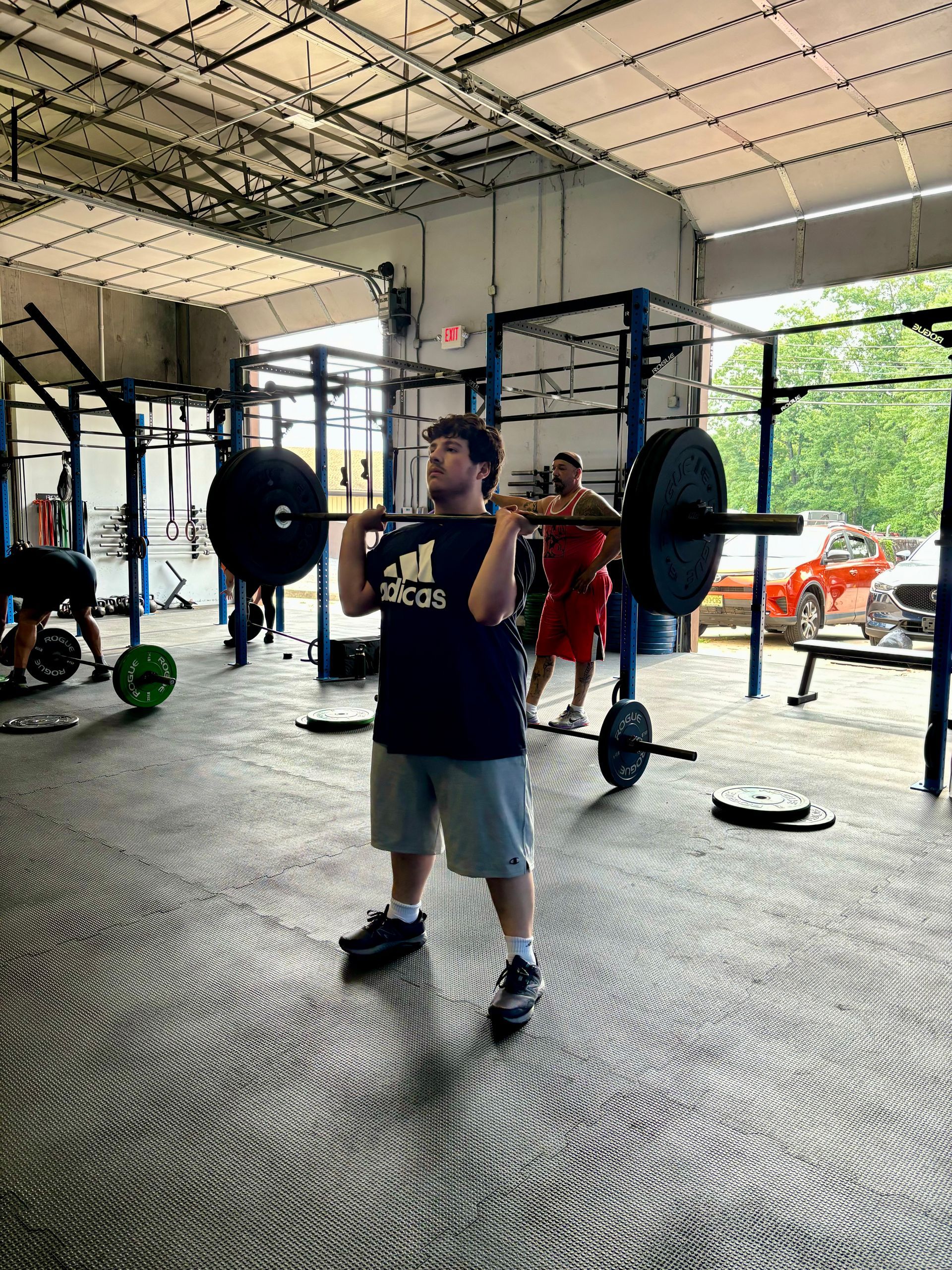 Man in gym lifting barbell overhead. He wears gray shorts and a dark shirt.