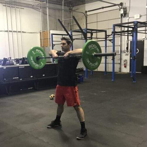 Man in gym holding barbell at shoulder level; green weights; red shorts.
