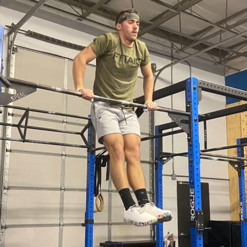 Man in gym, holding onto a pull-up bar, legs extended, wearing olive shirt, gray shorts, and colorful shoes.