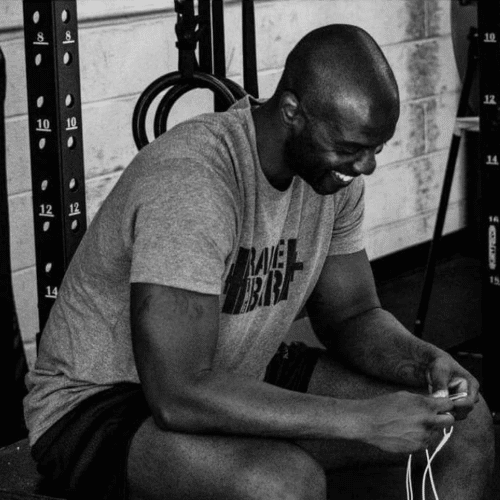 Man in a grey shirt smiling, sitting in a gym, holding earbuds.