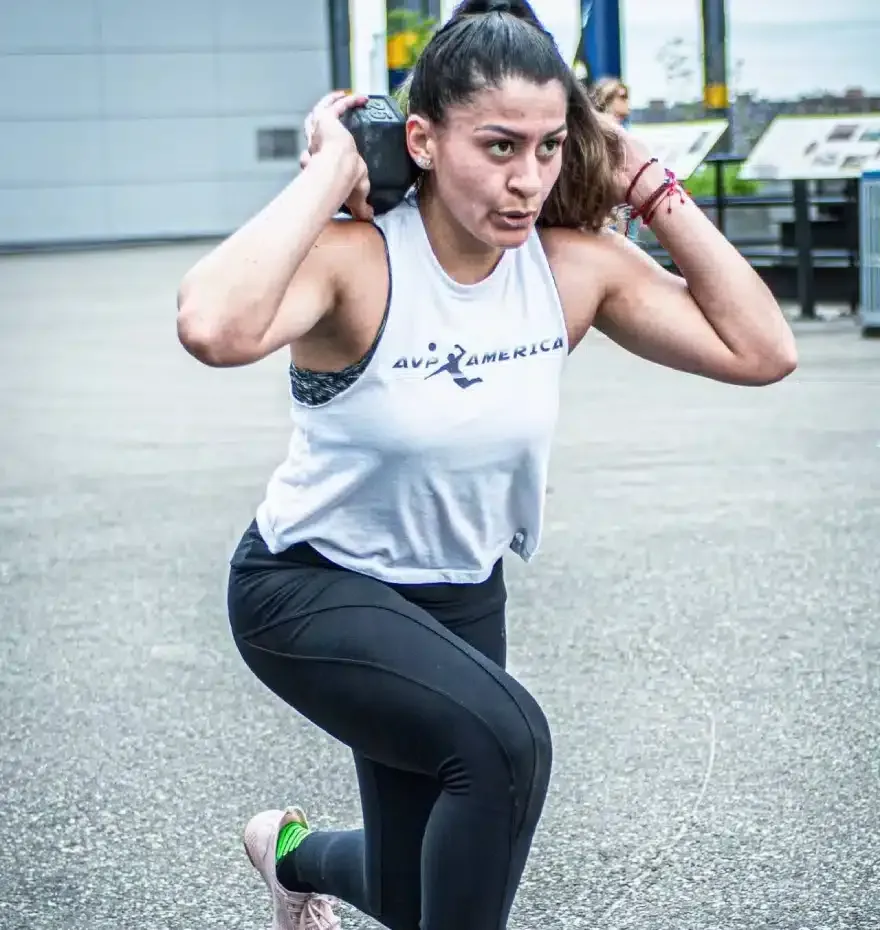Woman in workout gear doing a lunge with a weight held on her shoulders, outdoors.