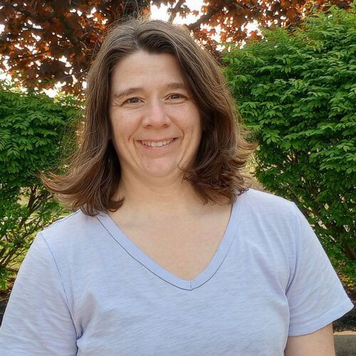Woman with brown hair smiles outdoors, wearing a light purple v-neck shirt, green bushes and tree behind her.