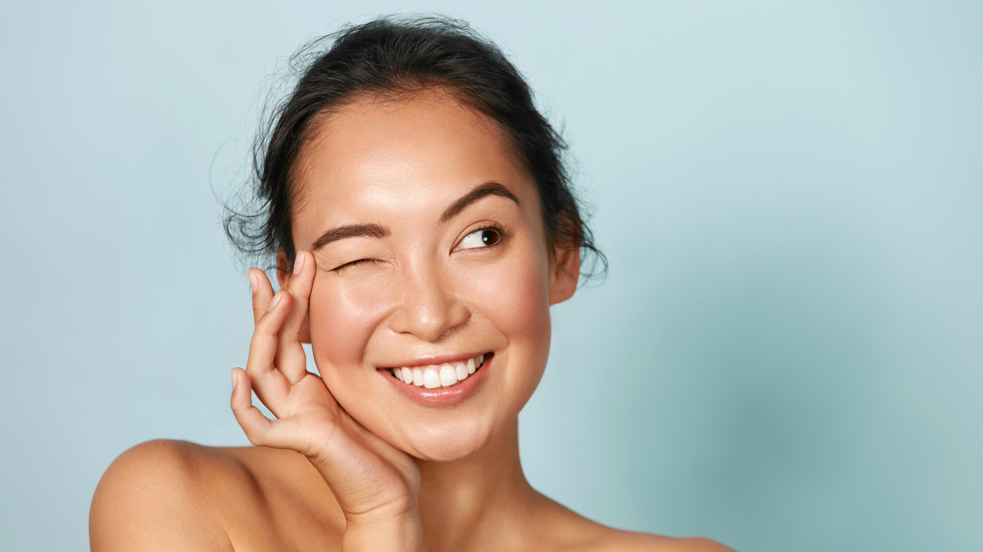 Woman with radiant skin winking and smiling, hand touching face, against a light blue background.