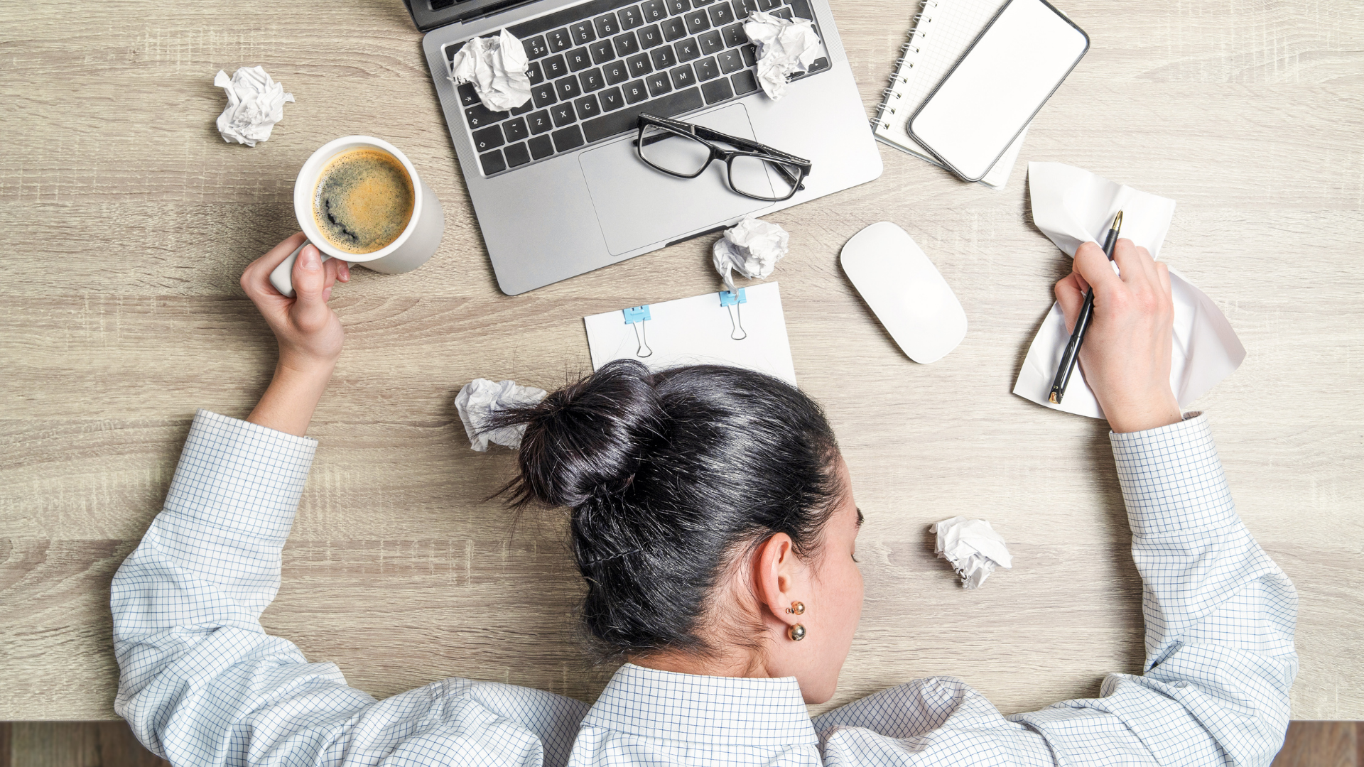 Woman asleep on a desk, surrounded by a laptop, coffee, crumpled paper, and a phone.