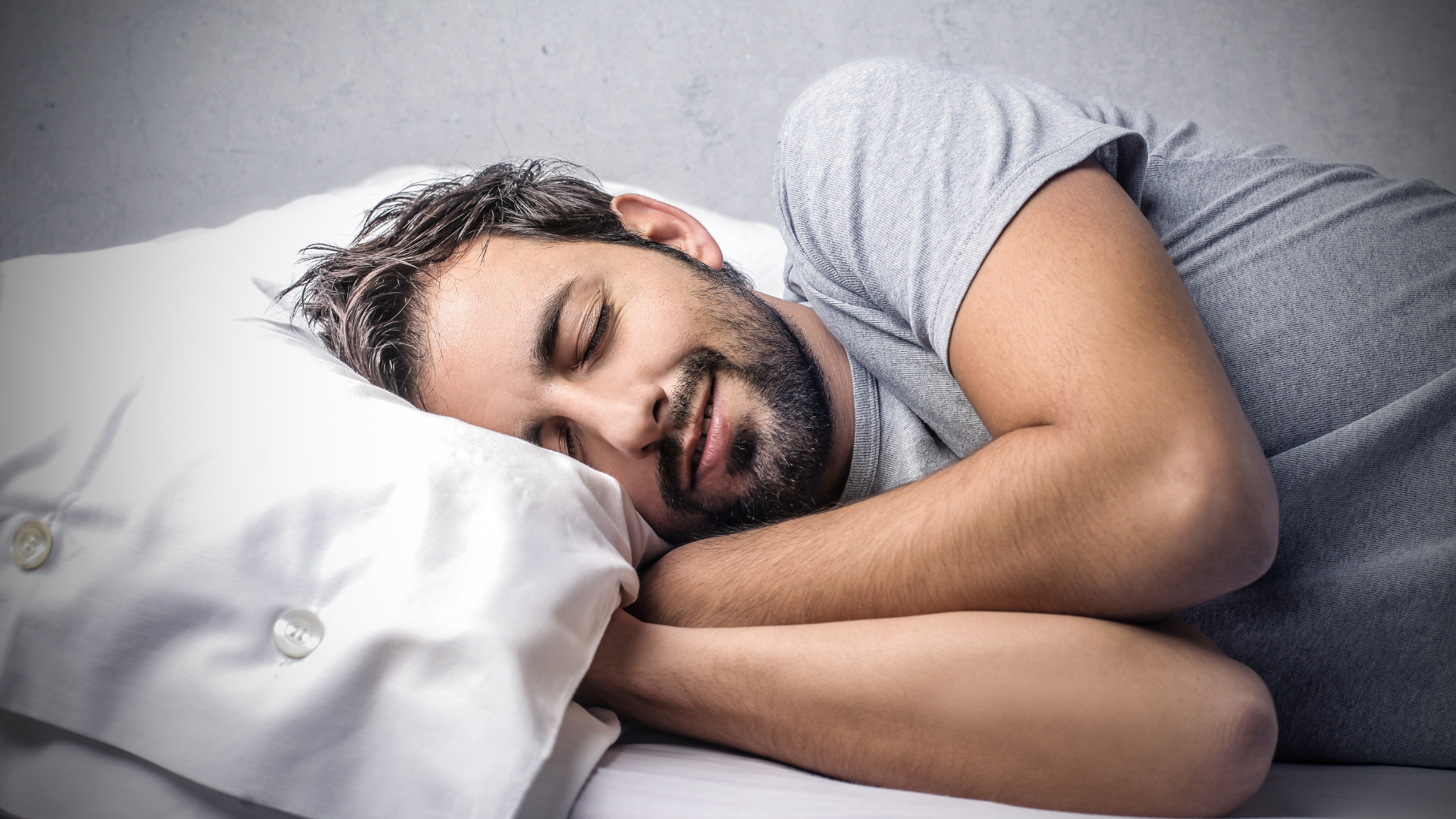 Man sleeping in bed, smiling, wearing a gray shirt, with a white pillow and sheet.
