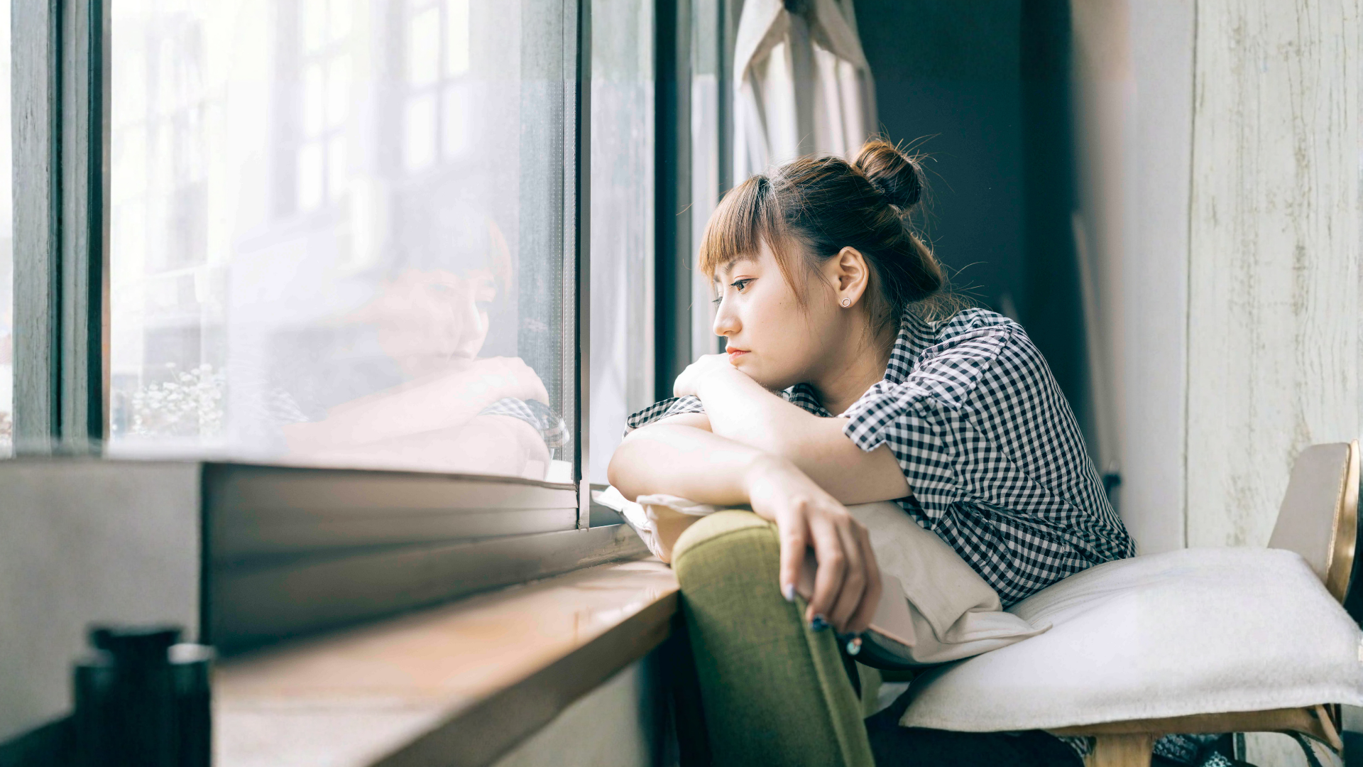 Woman resting chin on arm, looking out a window. Indoors, natural light.