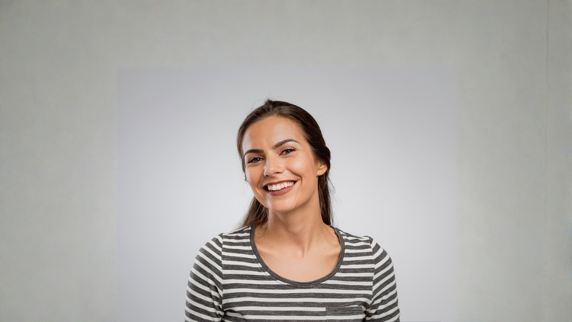 Woman with dark hair, smiling broadly, wearing a gray striped shirt, against a gray background.