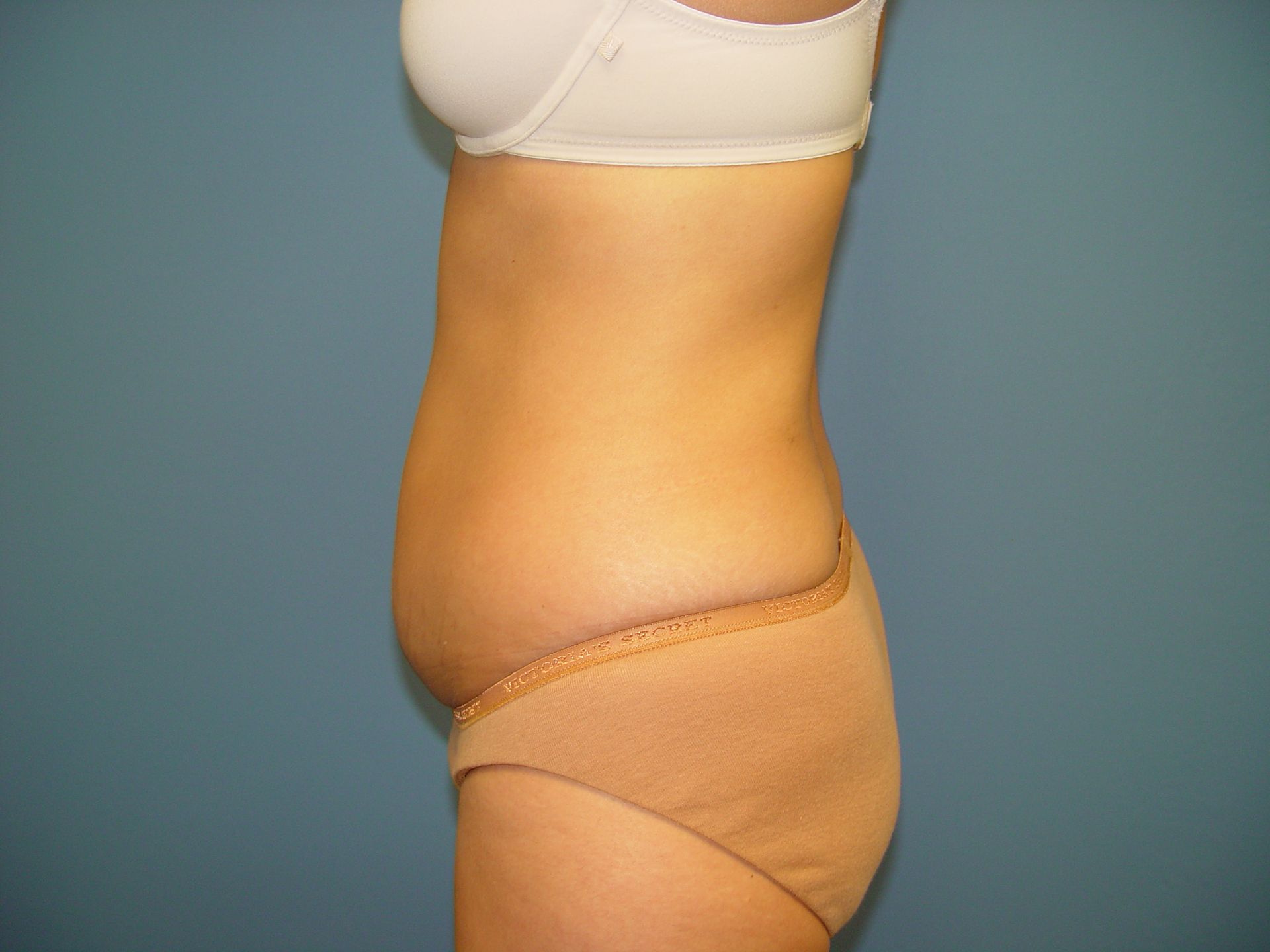 A woman in a white bra and tan underwear is standing in front of a blue wall.