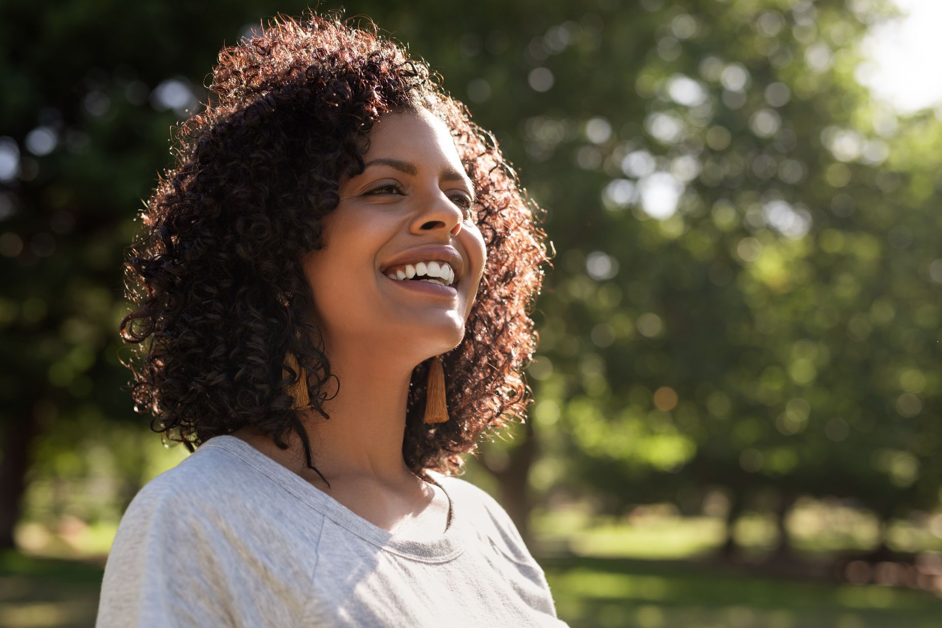 A woman with curly hair is smiling in a park.