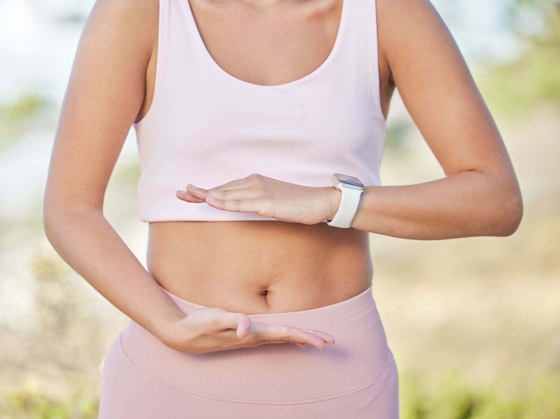 A woman in a white tank top and pink pants is holding her hands on her stomach.