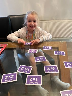 A young girl smiles while playing multiplication flash cards on a table. Cards are purple.