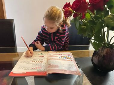 Girl writing in a workbook at a table, roses in vase nearby. She wears a striped shirt.