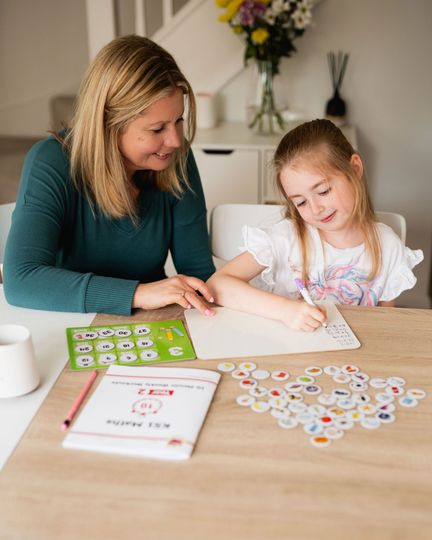 Sarah Gearing helping a child write on a whiteboard at a table. Colorful learning game and flowers present.