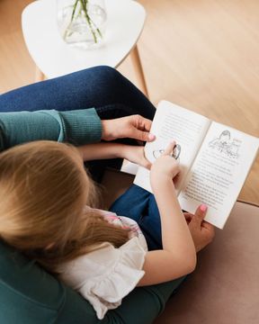 A child points at a book while sitting in a lap. Adult in teal sweater, white top and blue jeans.