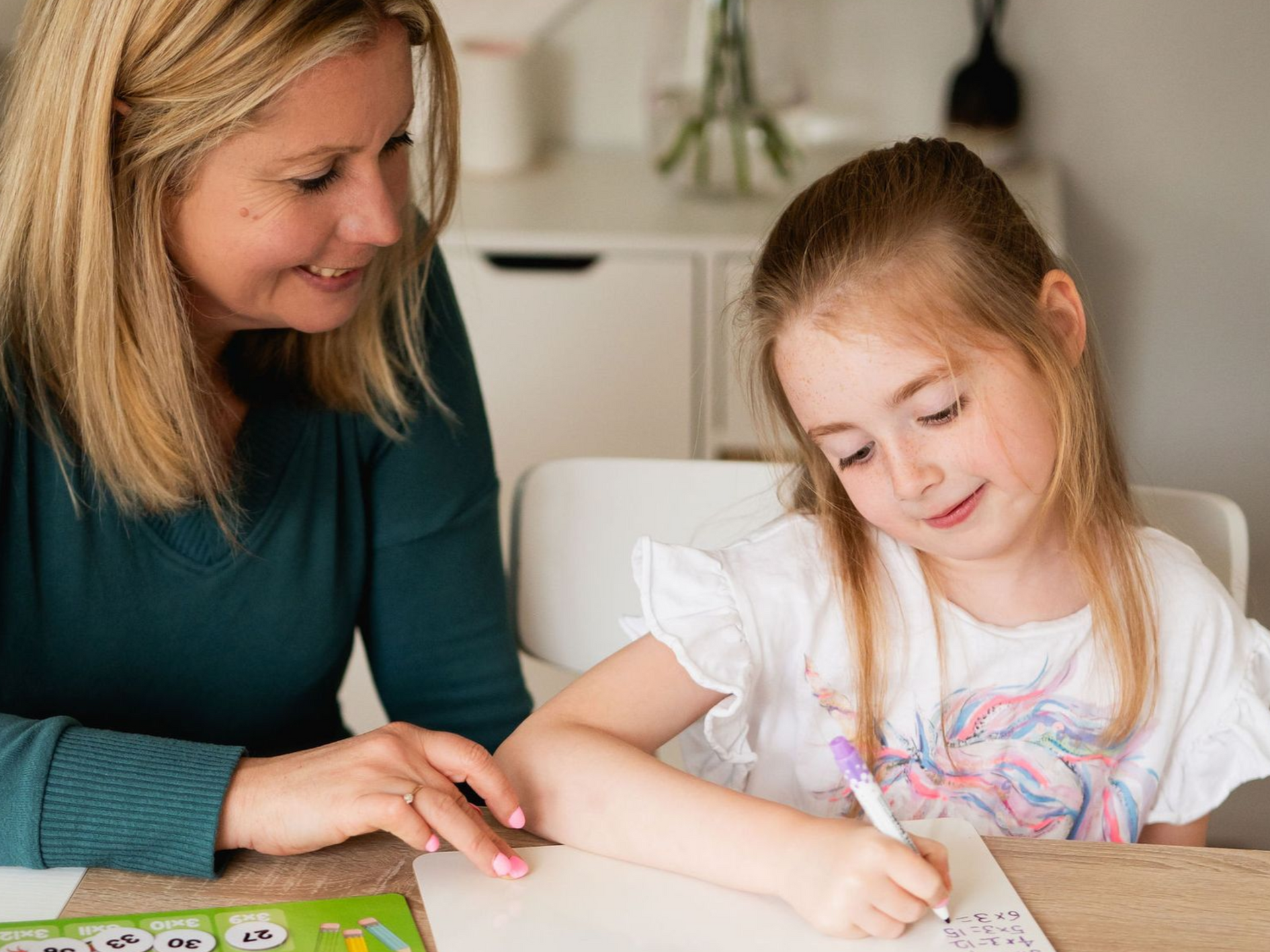 An adult and child sit at a table, with the child writing on paper and the adult watching attentively.