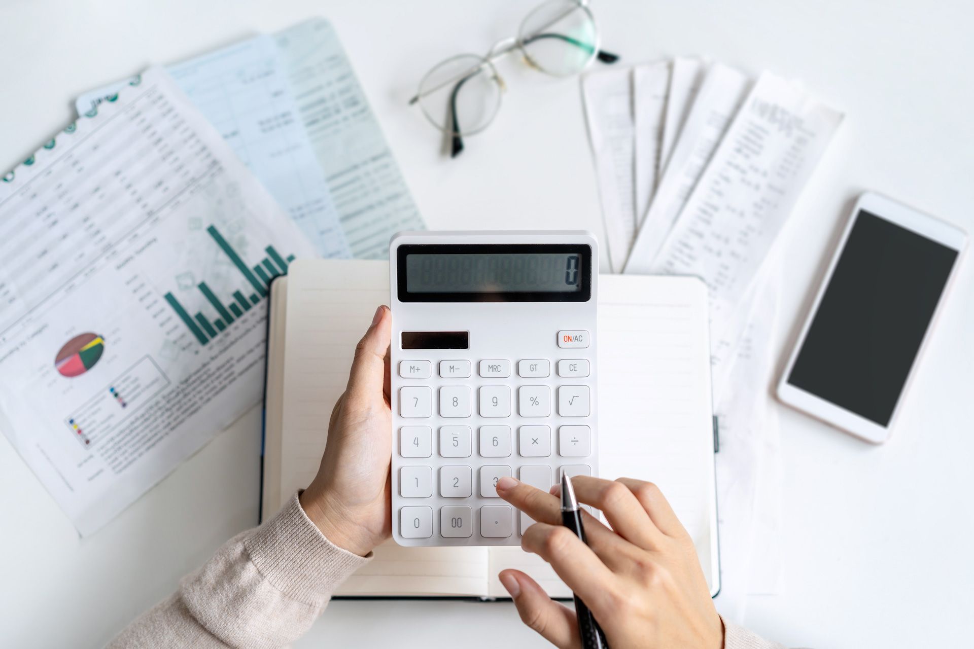 A person is using a calculator on a desk with papers and a cell phone.