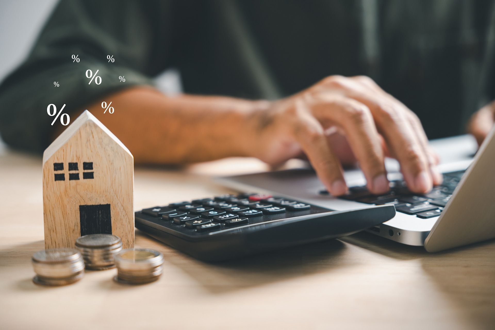 A man is typing on a laptop next to a calculator and a model house.