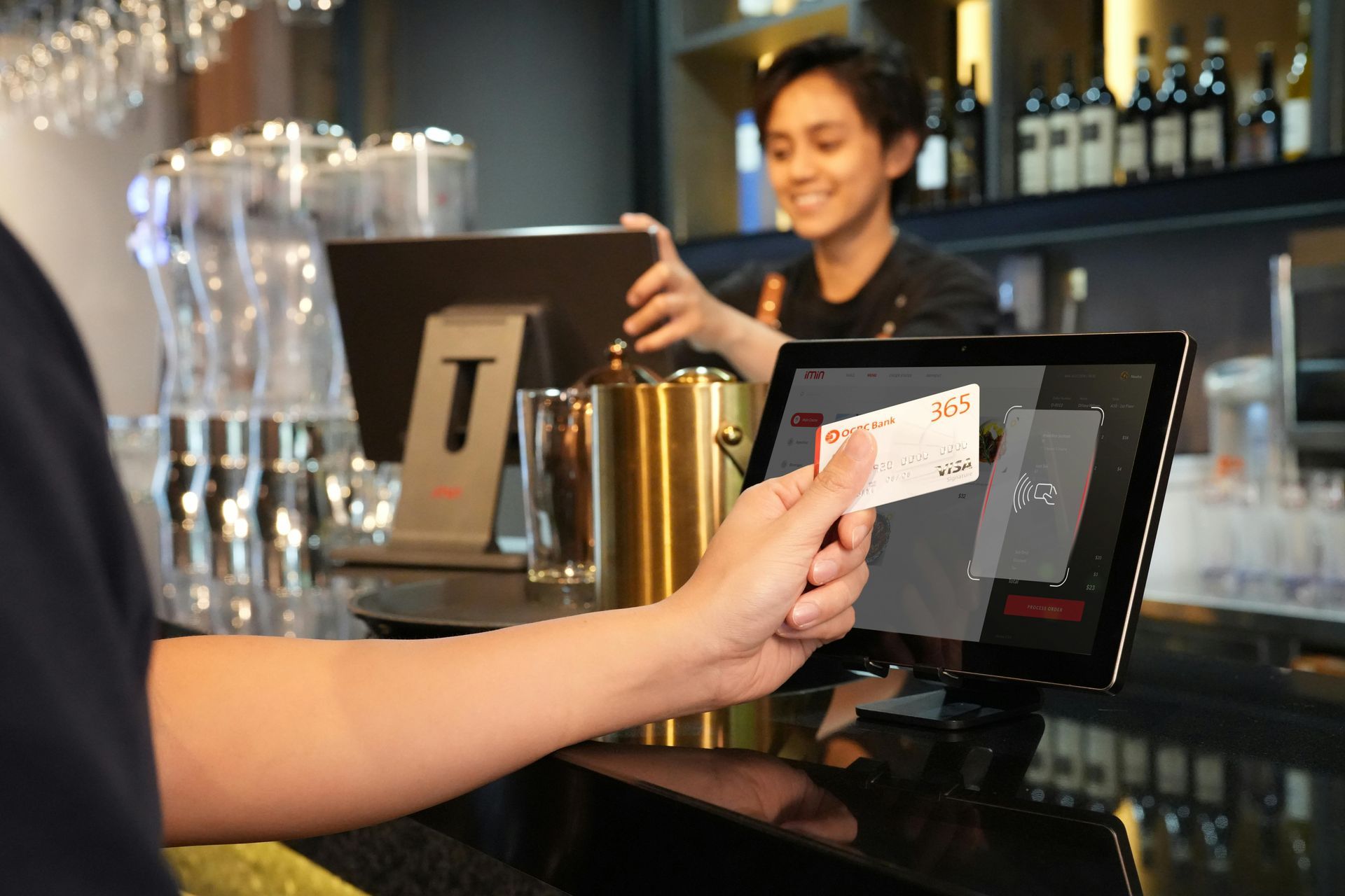 A customer uses a contactless payment card at a terminal at a bar counter while an employee smiles in the background.