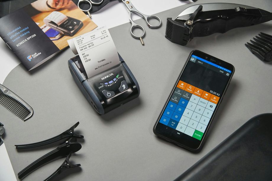 A portable thermal printer, a smartphone showing a POS app, scissors, and hair clipper accessories on a gray surface.