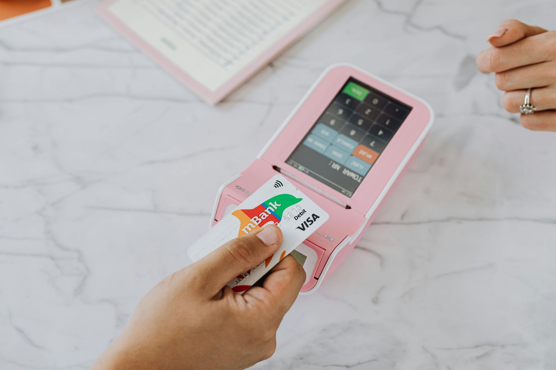A hand holds a credit card over a pink contactless payment terminal on a white marble surface.