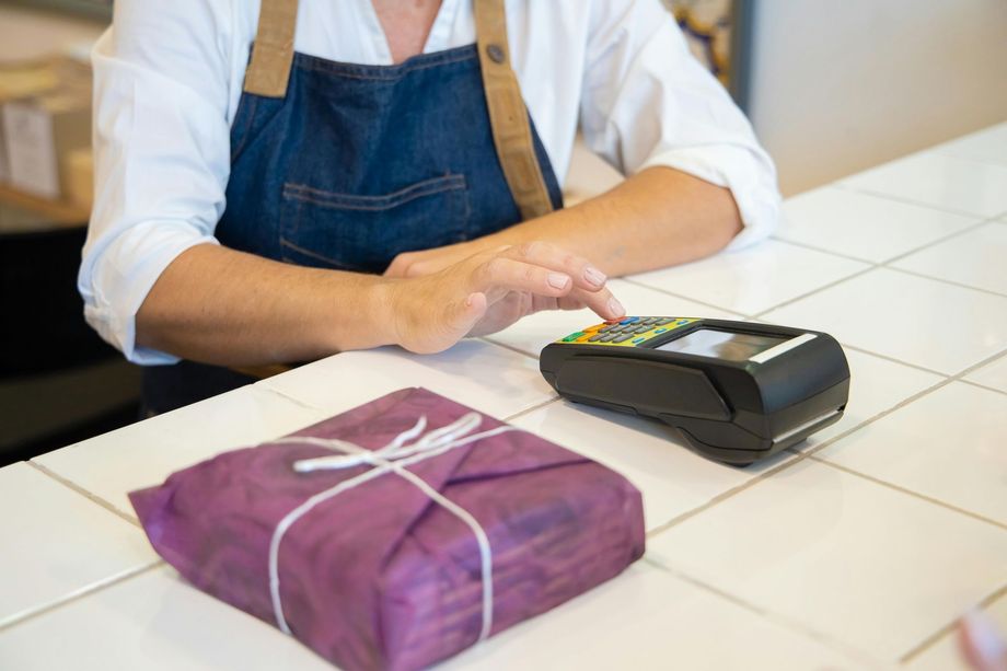 A person in a blue apron processing a payment on a card reader next to a purple-wrapped package on a tiled counter.