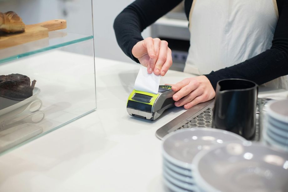 A person behind a cafe counter tears a printed receipt from a handheld payment terminal.