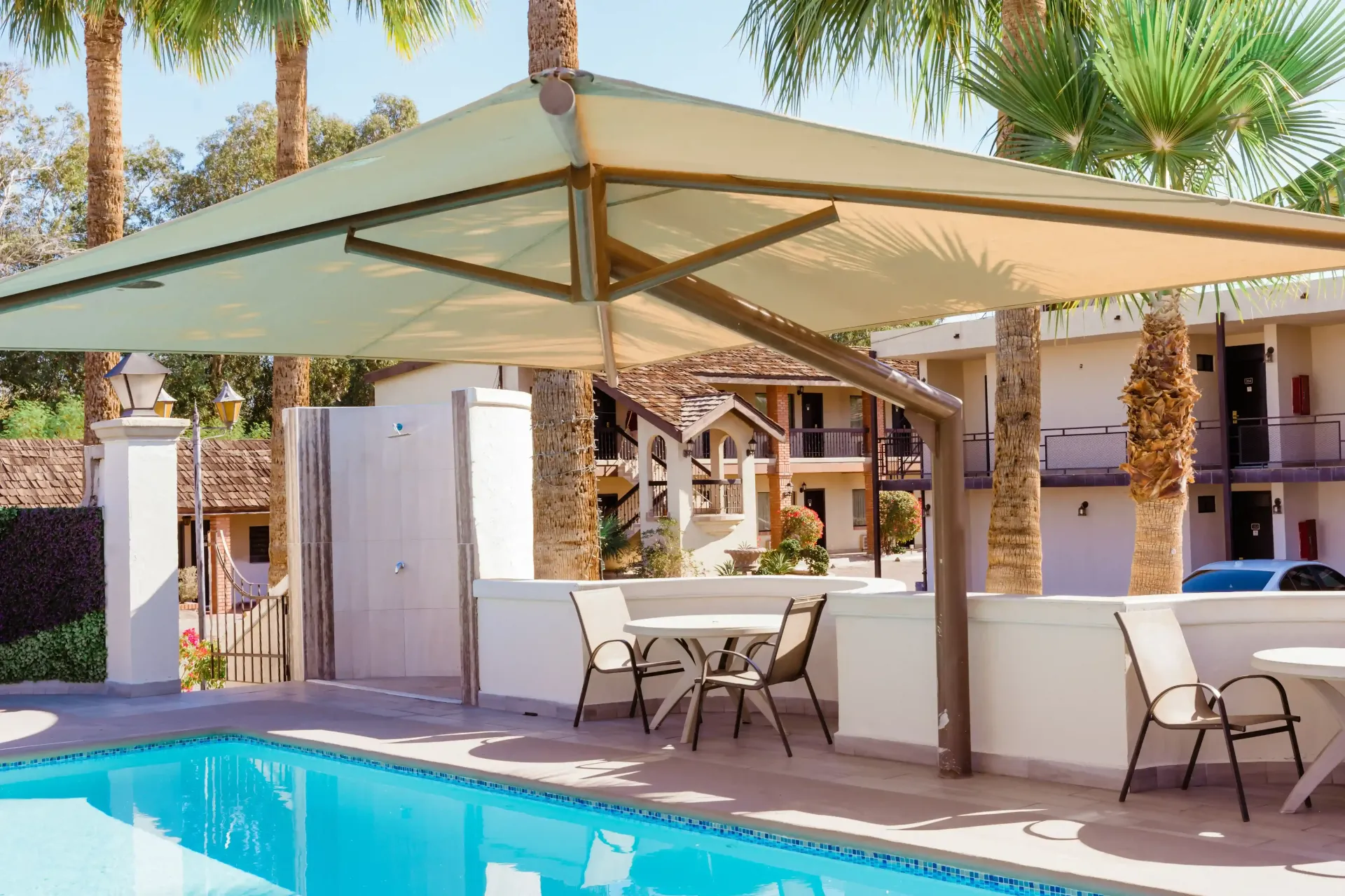 Complejo turístico, una sombrilla voladiza beige da sombra a un patio junto a la piscina con sillas, mesas y palmeras.