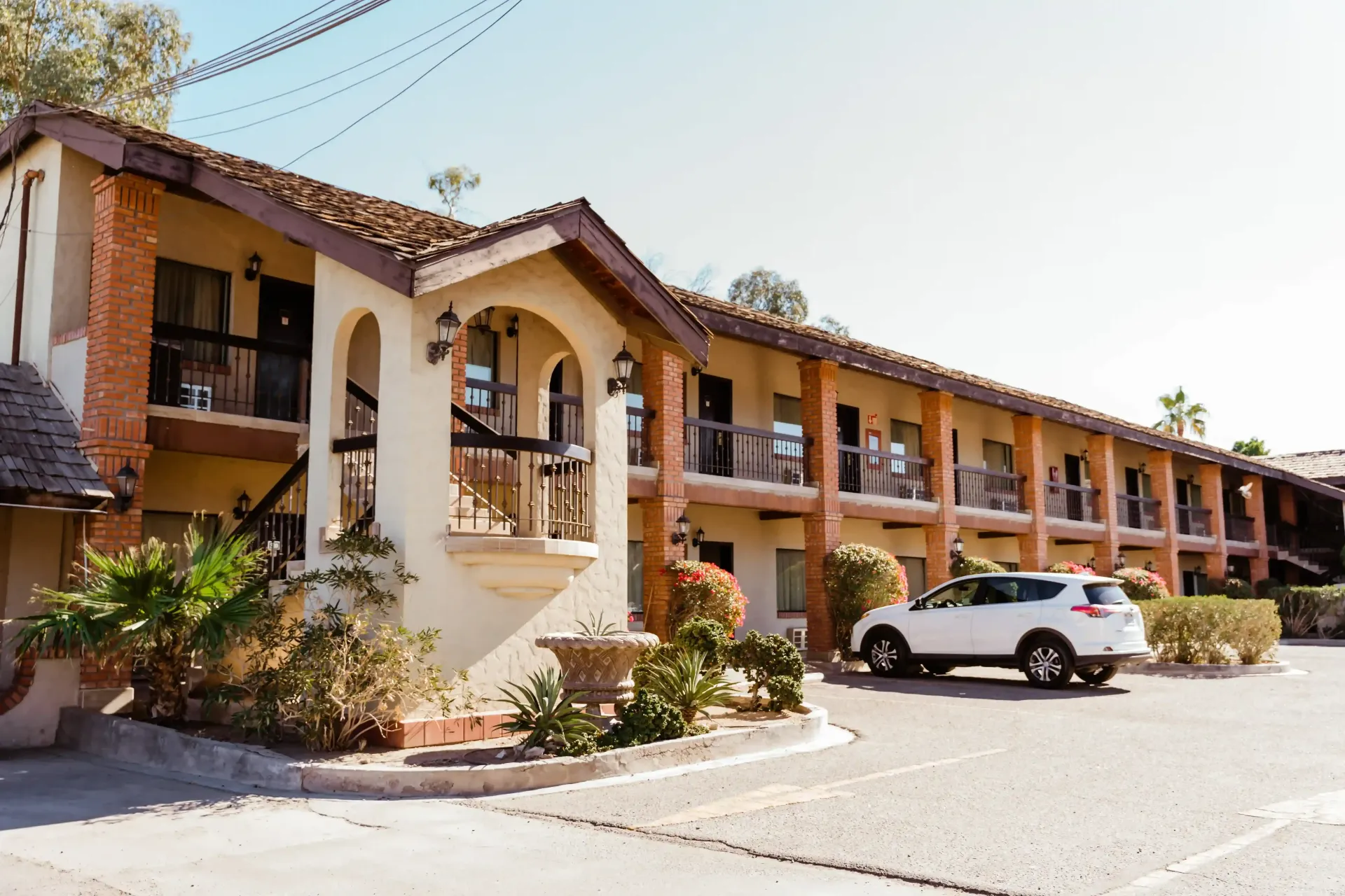 Hotel de dos pisos con fachada de estuco, tejado marrón y entrada arqueada, con un coche blanco aparcado delante.