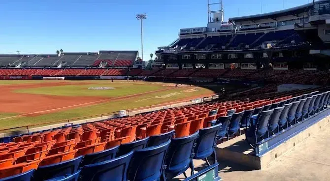 Filas de asientos vacíos en un estadio de béisbol con un campo al fondo.