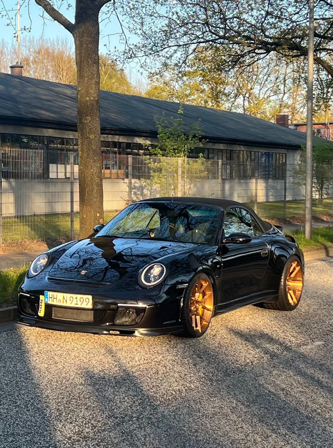 Schwarzes Porsche-Cabrio mit goldenen Felgen, an einem sonnigen Tag auf einer Straße vor einem Gebäude geparkt.