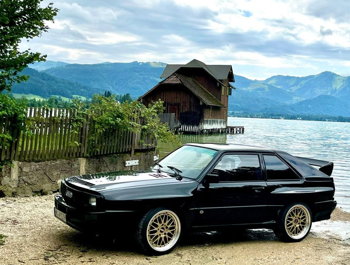 Schwarzes Audi Quattro Coupé, geparkt an einem Strand am See, mit hölzernem Bootshaus und Bergkulisse.