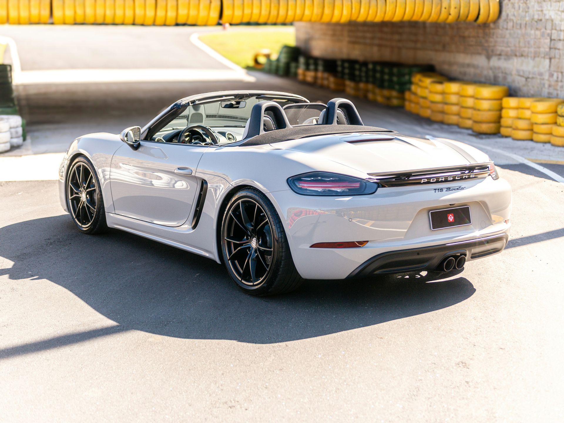 Ein hellgraues Porsche 718 Boxster Cabriolet parkt auf einer Rennstrecke; im Hintergrund sind gelbe Leitplanken zu sehen.
