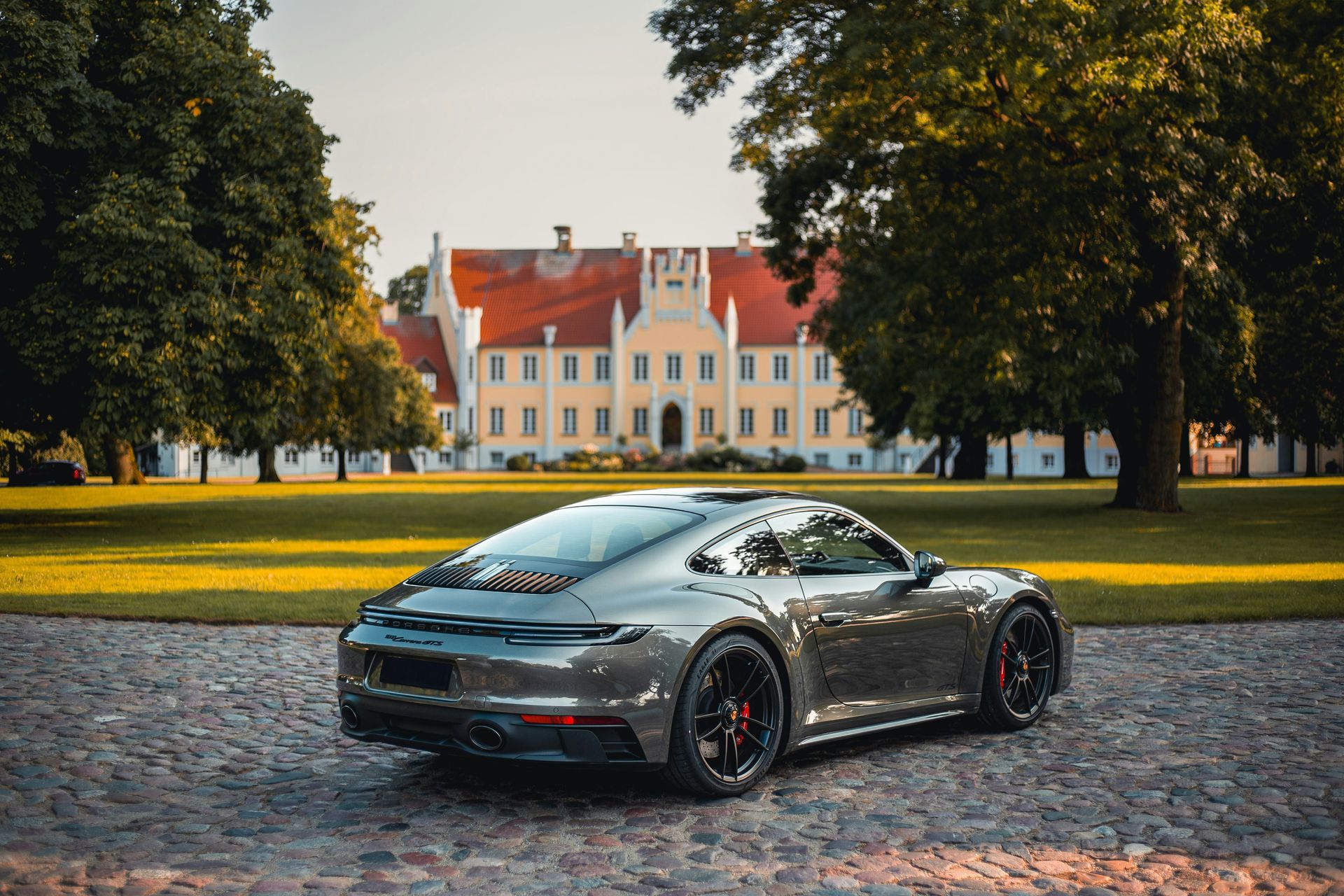 Ein dunkelmetallischer Porsche-Sportwagen parkt auf einem Kopfsteinpflasterweg vor einem Herrenhaus mit rotem Dach.