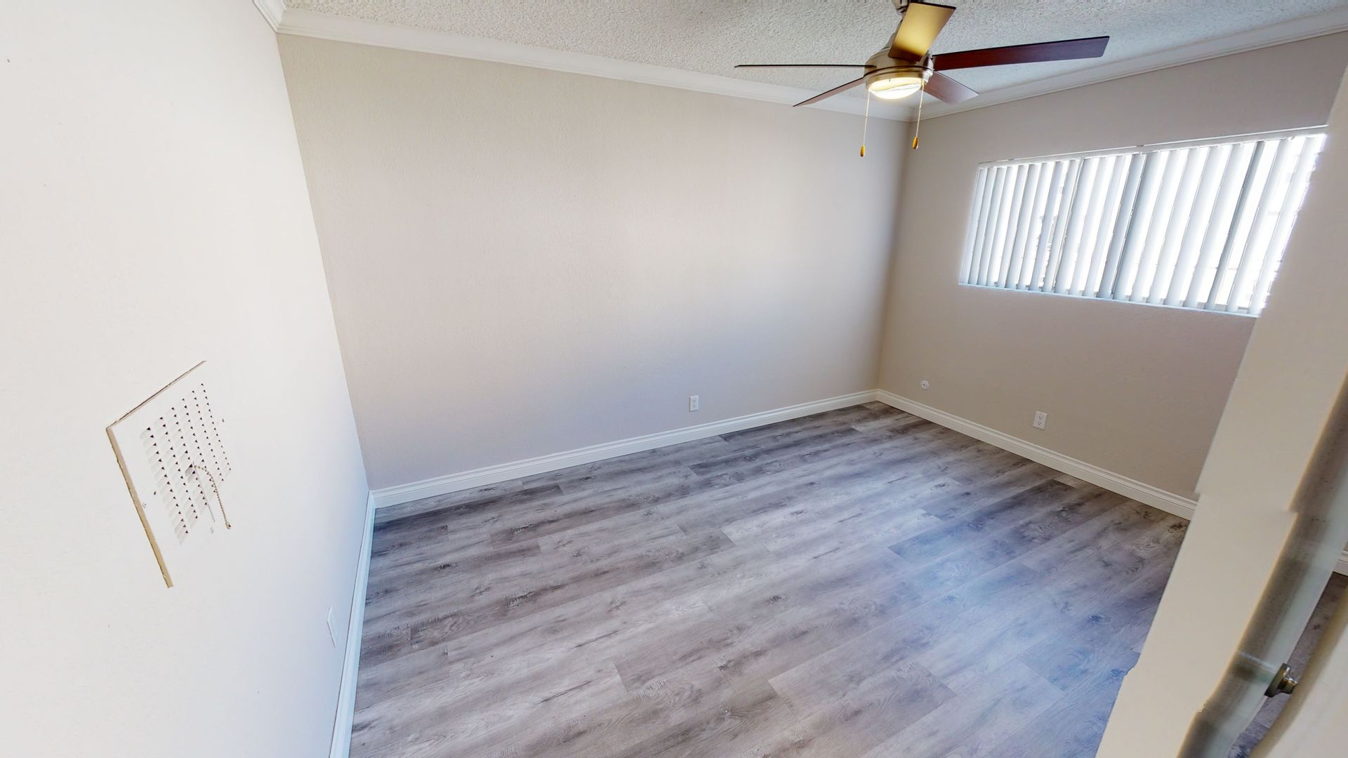 Empty bedroom with grey wood-look flooring, beige walls, and window with blinds. Ceiling fan present.