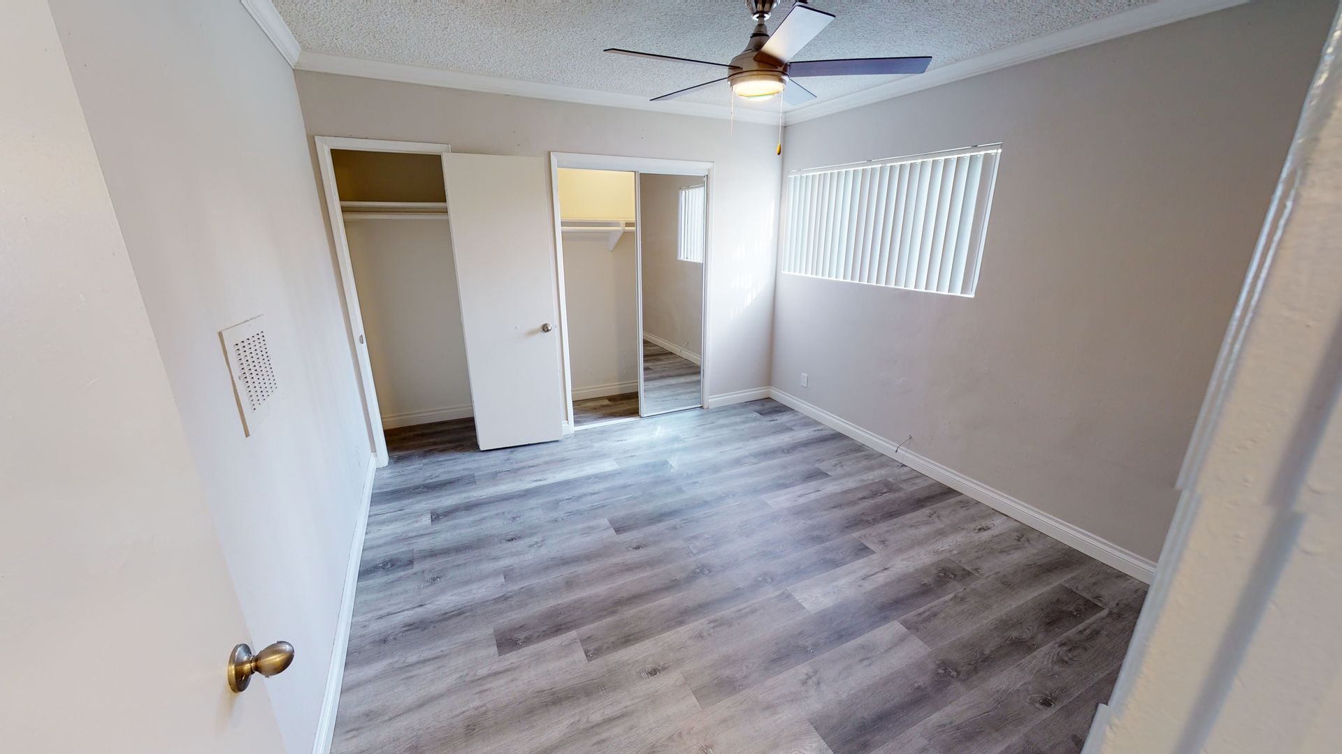 Empty bedroom with gray wood-look flooring, ceiling fan, open closet, and window with blinds.