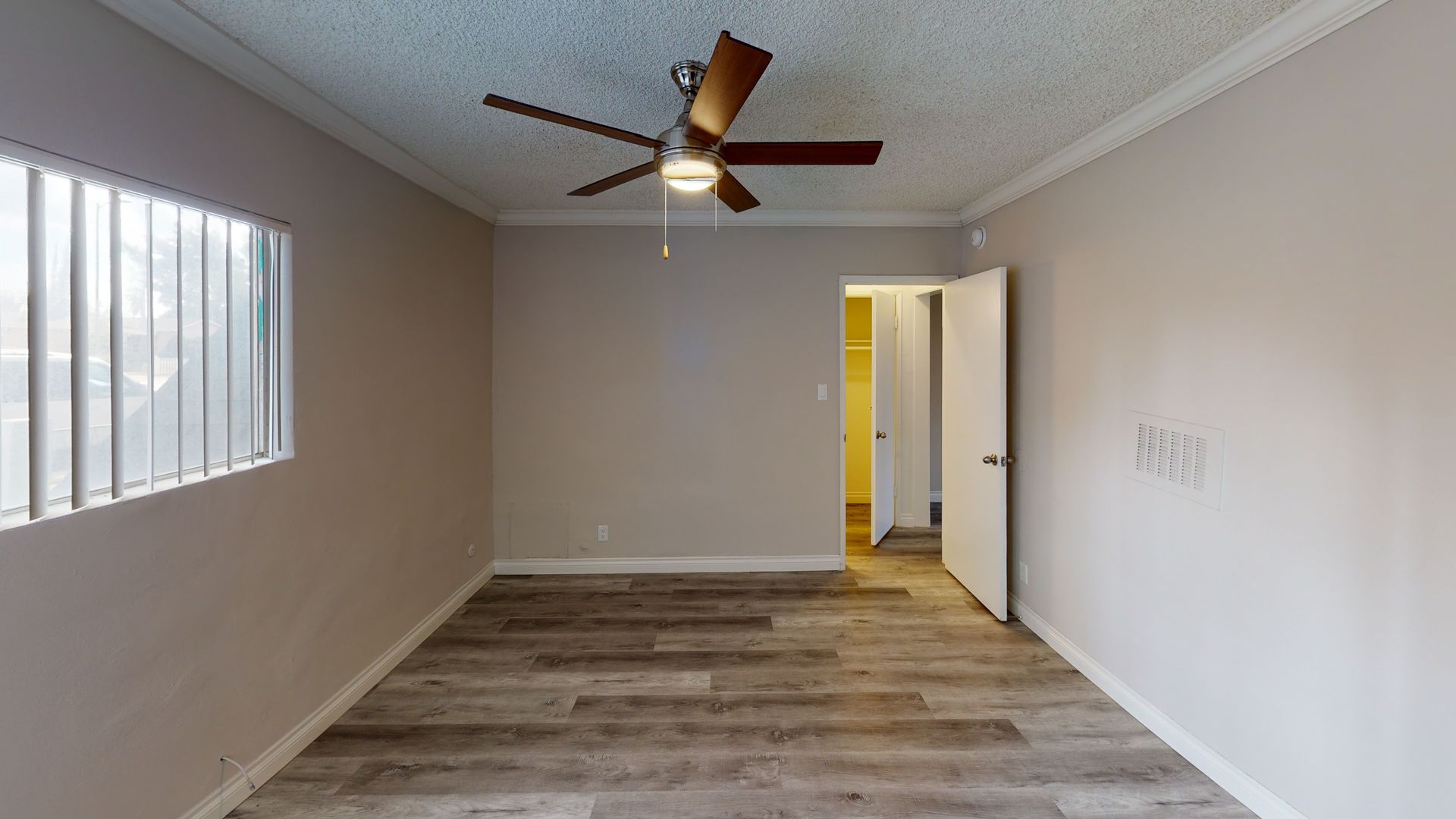 Empty bedroom with wood-look flooring, tan walls, ceiling fan, open door, and a window with blinds.