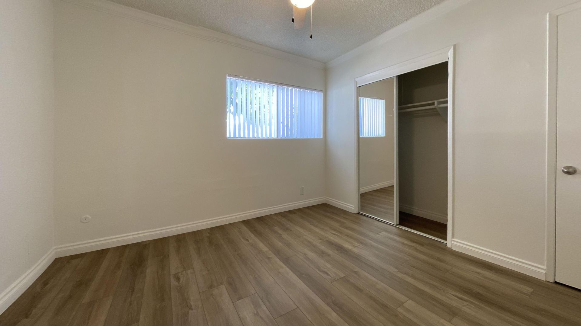 Empty bedroom with wooden floor, closet, window, and white walls.