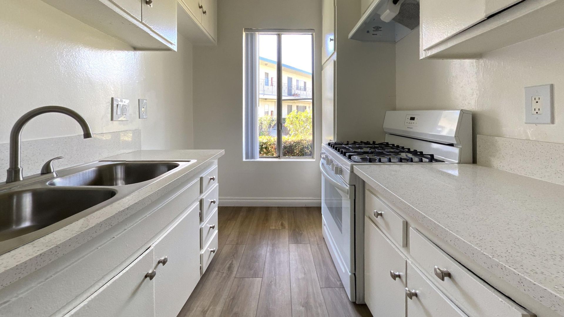 A narrow white kitchen with a window, stove, and sink. White cabinets, wood-look floor.