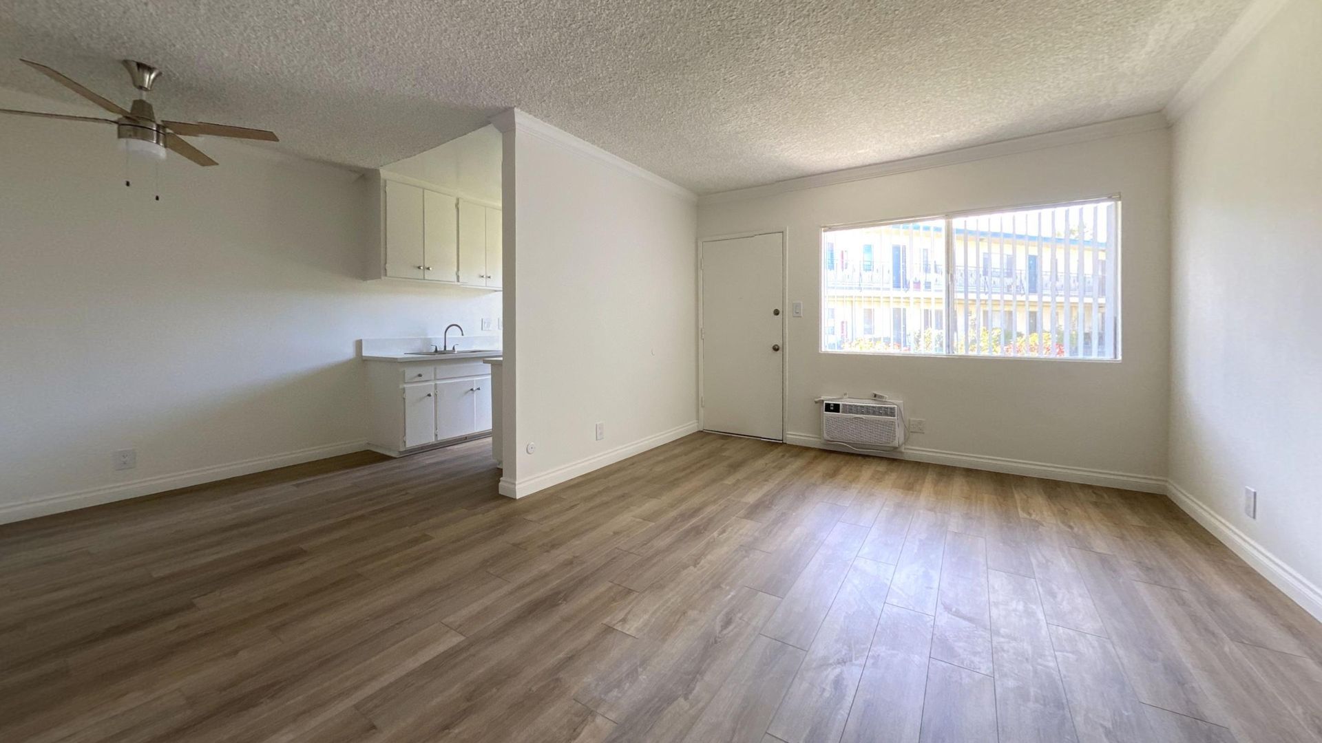 Empty apartment room with wood flooring, white walls, and a small kitchen visible.